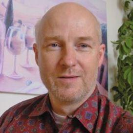 Mann mit kurzem, leichtem Bart, im gemusterten Hemd, vor einem Weingemälde und einer Pflanze.